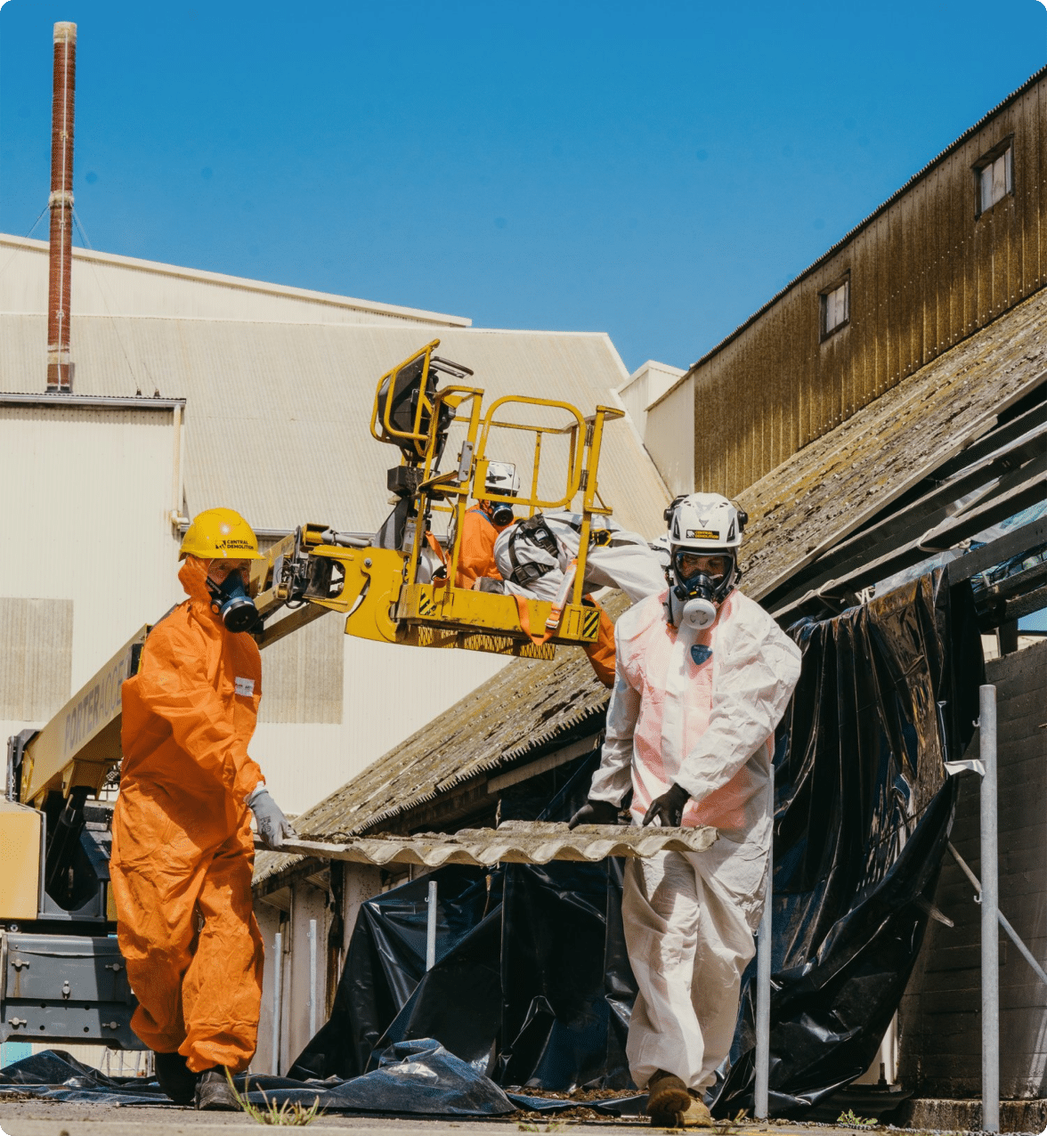 Two workers in protective gear remove roofing material containing asbestos.