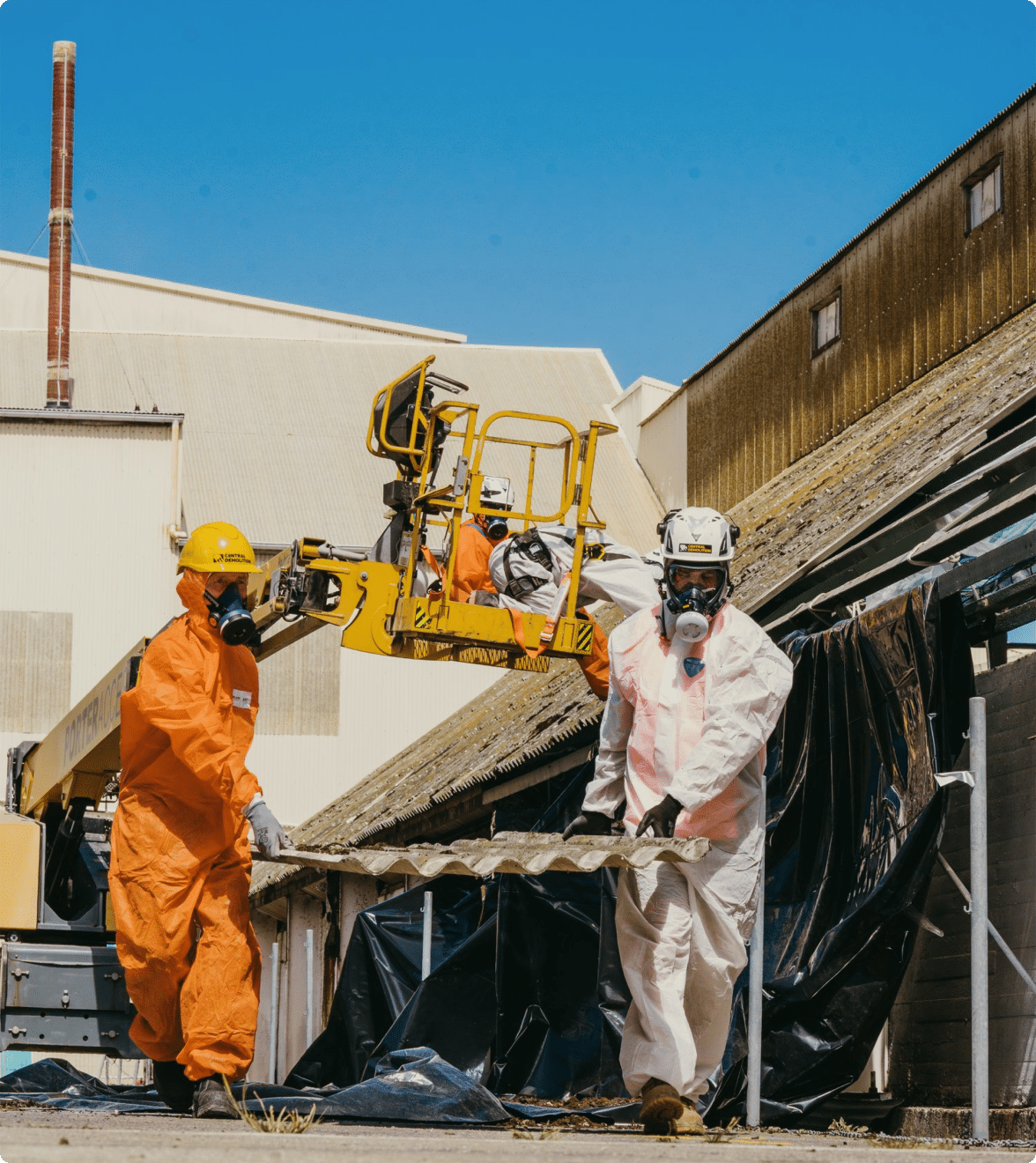 Two workers in protective gear remove roofing material containing asbestos.